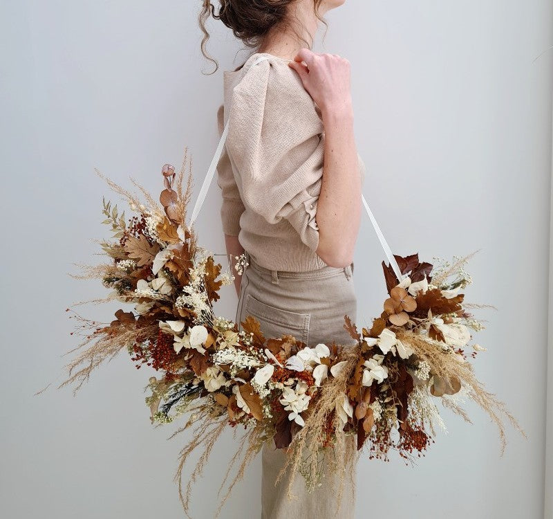 Vrouw die een moderne bloemenkrans in de vorm van een halve maan met droogbloemen zoals pampas en eikenblad in bruine en beige tinten met een lint aan haar schouders heeft hangen