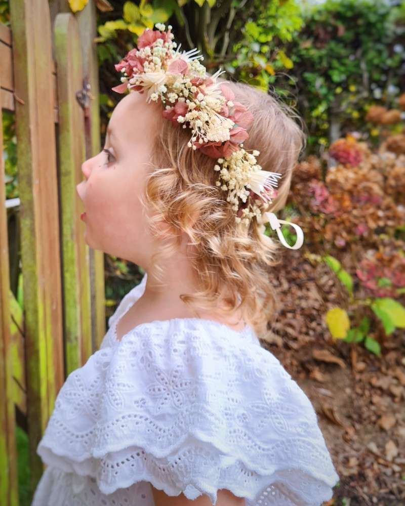 communiefoto van meisje met krullen die over een hek kijkt, een wit zwierig kleedje draagt en een weelderige bloemenkroon draagt gemaakt van droogbloemen zoals paarse hortensia, gipskruid en eucalyptus