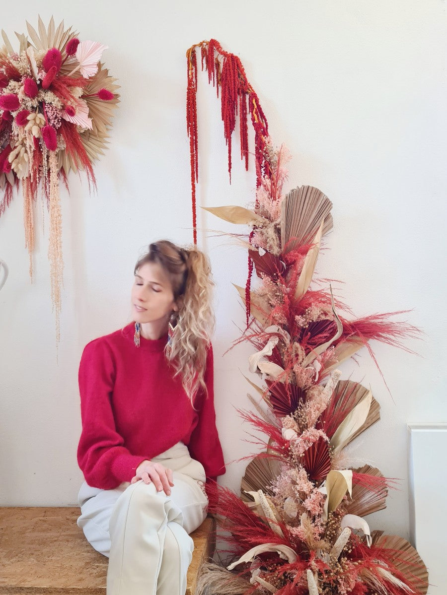 Jonge vrouw met rode trui die op een bank zit naast een droogbloemeninstallatie die tegen de muur werd gemaakt met gedroogd palmblad, amaranthus en pampas in rode en beige tinten