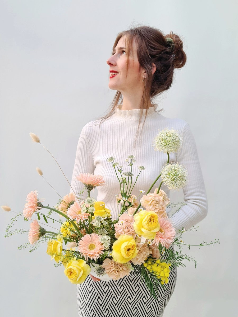 Jonge vrouw die modern bloemstuk voor zich houdt gevuld met verse snijbloemen zoals alium, zalmroze anjers, mimosa in een basic witte schaal