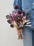 Vrouw die gebonden boeket droogbloemen vasthoudt in overwegend groene tinten meteen vleugje wit en paars, met eucalyptus, echinops, broom, palmspeer en jazilda