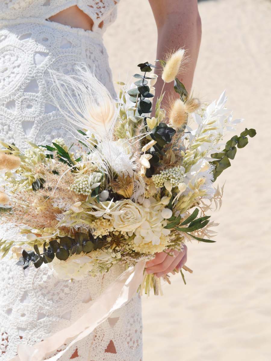 close-up van een modern bruidsboeket van droogbloemen in witte en groene tinten, door een bruid langs haar zijde gehouden