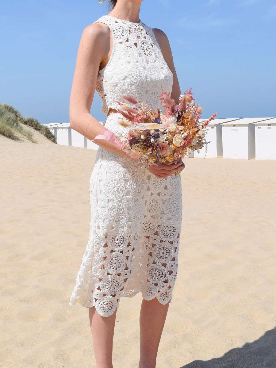 Bruid staat op het strand tussen de duinen en draagt een bruidsboeket van droogbloemen met roze bunny tails, eucalyptus, witte gepreserveerde rozen en tarwe.
