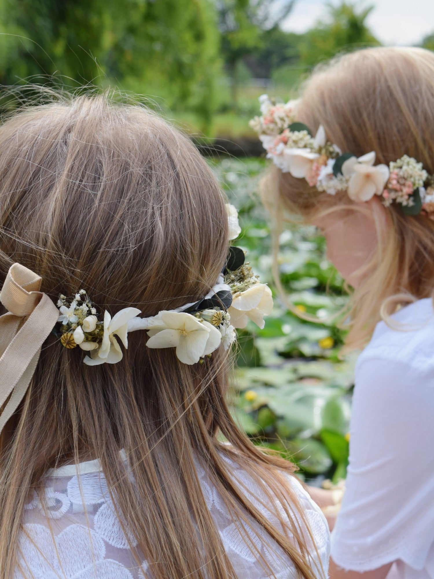 Twee meisjes zitten aan een vijver en dragen een zwierig wit kleedje en een bloemenkrans gemaakt van droogbloemen in zachtroze en witte witten