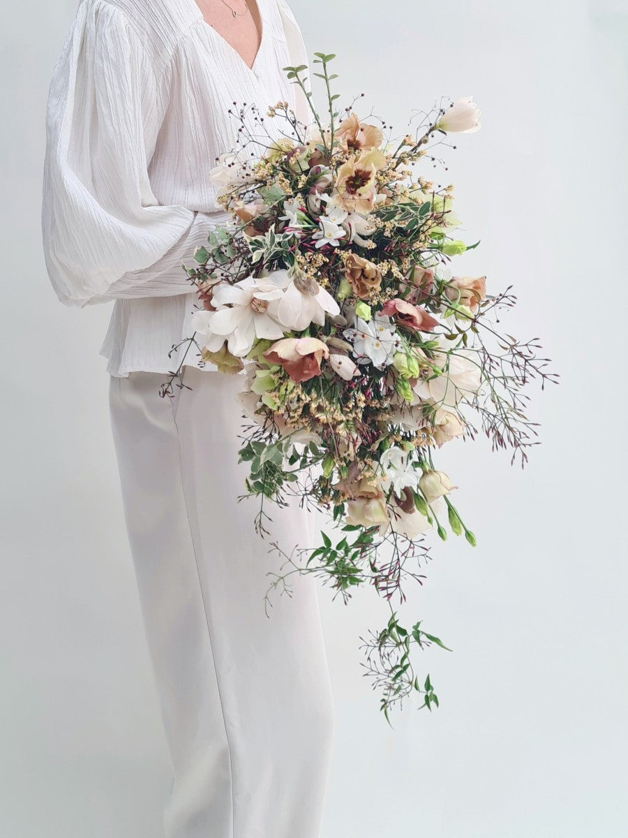 Vrouw met witte outfit heeft modern cascade bruidsboeket in de hand met afhangende bloemen zoals mokka lysianthus, viburnum, magnolia, jasmina in bruine en roze tinten