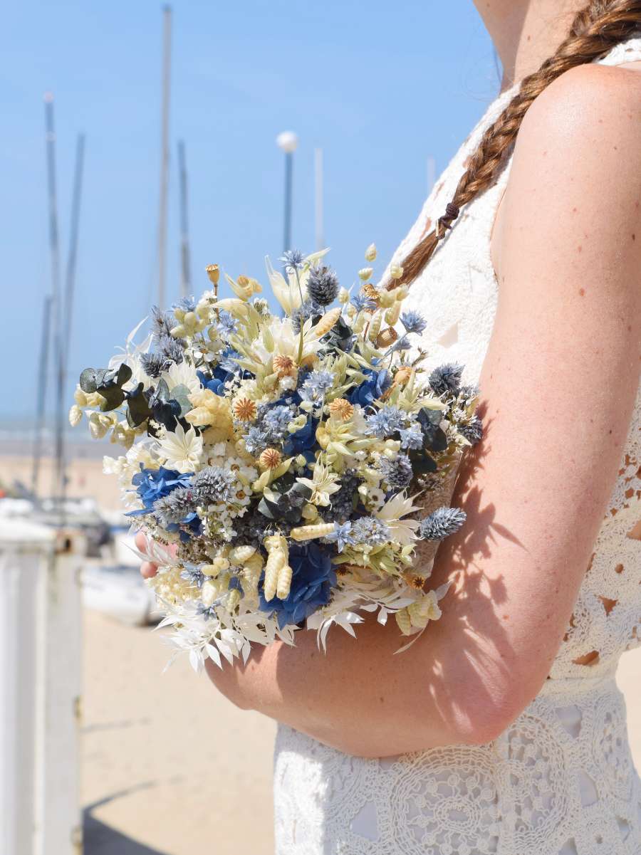 Bruid met wit kleedje houdt rond en compact bruidsboeket van droogbloemen vast in groene, blauwe en witte tinten. Op de achtergrond zijn de zee en boten te zien.