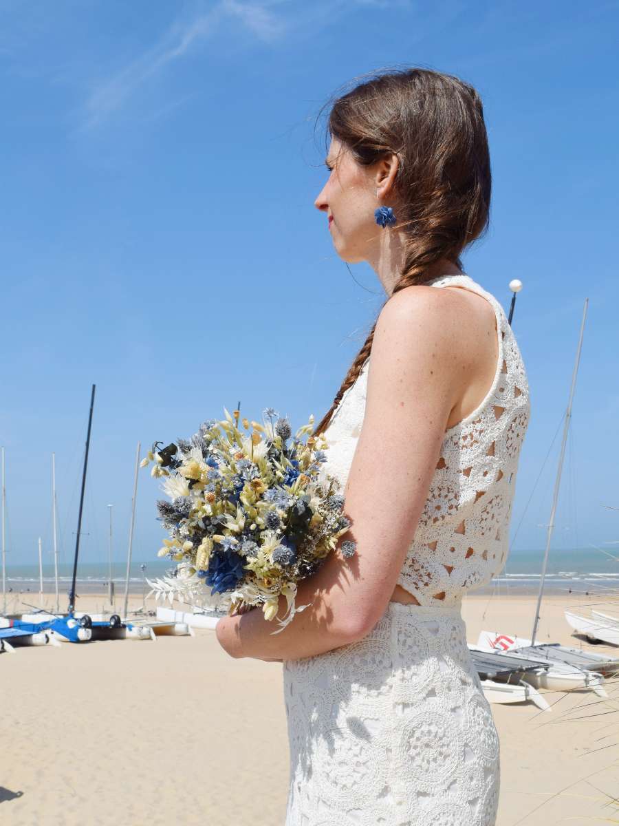 Bruid kijkt naar de zee en draagt in haar armen een bruidsboeket van droogbloemen in een ronde vorm met blauwe, groene en witte tinten. Ze draagt bijpassende blauwe oorbellen van droogbloemen gemaakt