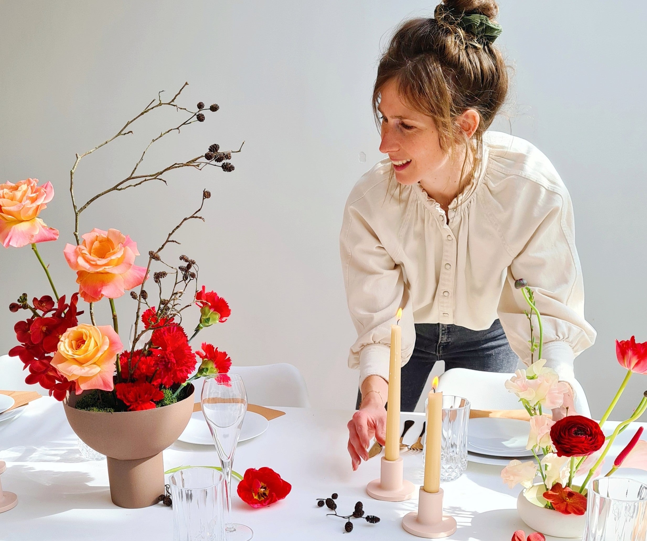 Aangeklede feesttafel met een modern ikebana bloemstuk, kaarsenhouders met lange kaarsen en een bloemiste die zich over de tafel heen buigt