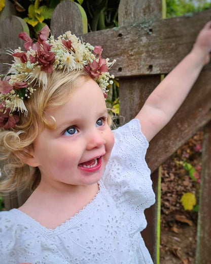communiefoto van klein meisje met krullen die een weelderige bloemenkroon met paarse hortensia, witte nigella en gipskruid draagt