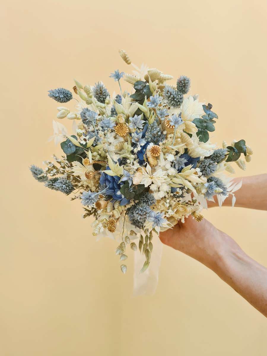 Close-up van een bruidsboeket gemaakt van droogbloemen in blauwe, grijze, witte en groene tinten, met een lang beige lint aan