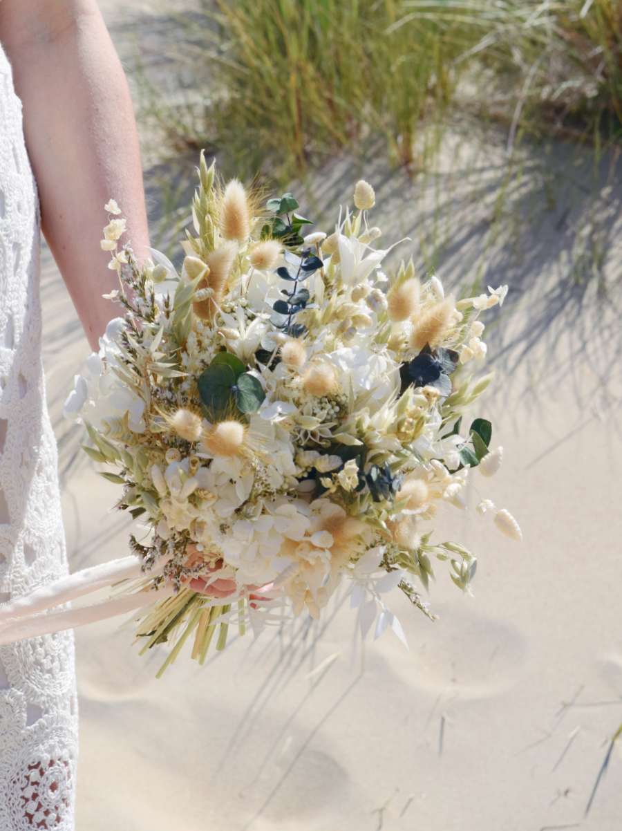 Bruid poseert aan de duinen met een modern en compact bruidsboeket van droogbloemen. Het boeket bevat groene eucalyptus, bunny tails, hortensia en andere witte droogbloemen
