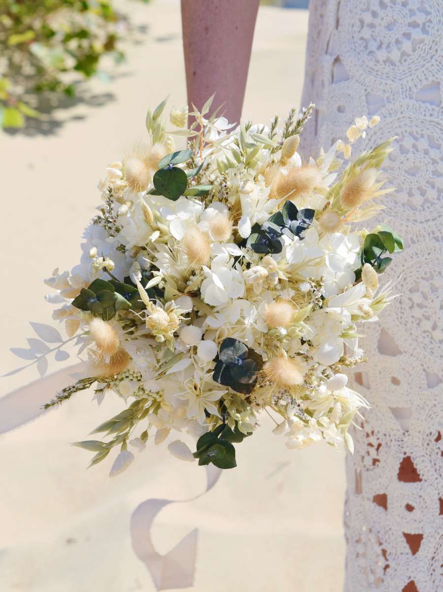 Bruid houdt een klein rond bruidsboeket van droogbloemen naast zich, opgemaakt met witte en groene droogbloemen zoals eucalyptus, hortensia en ruscus
