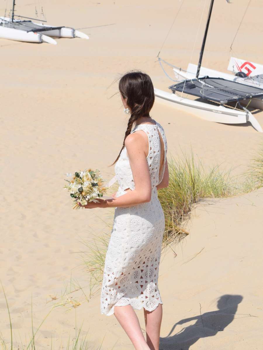 Bruid loopt de duinen af met een klein bruidsboeket van droogbloemen in haar hand, opgemaakt met groene en witte droogbloemen
