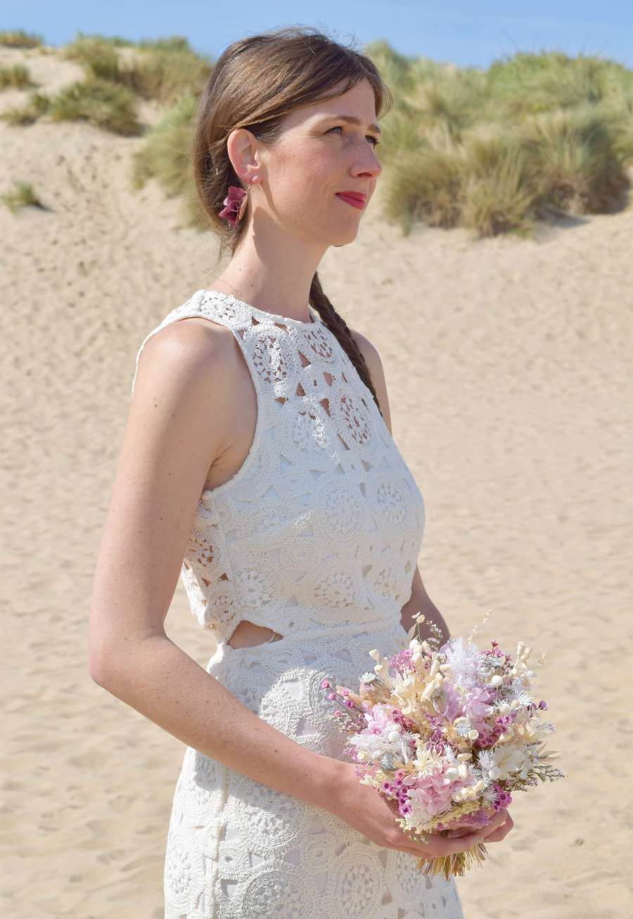 Bruid poseert aan de duinen met een klein bruidsboeket van droogbloemen in frisse pasteltinten waaronder lila hortensia, papavers, witte tatarica en glixia