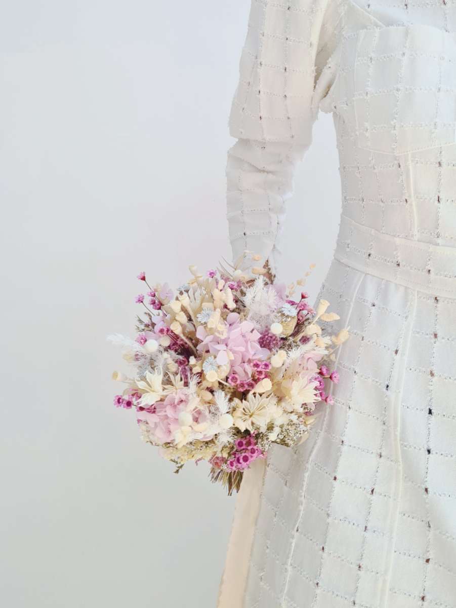 Model houdt een rond en compact trouwboeket van droogbloemen langs haar lichaam met frisse witte en lila tinten