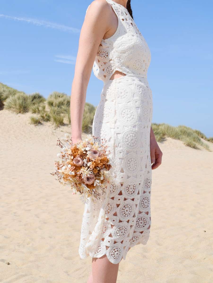 bruid poseert in de duinen met een klein trouwboeket van moderne droogbloemen in zachte mokktinten
