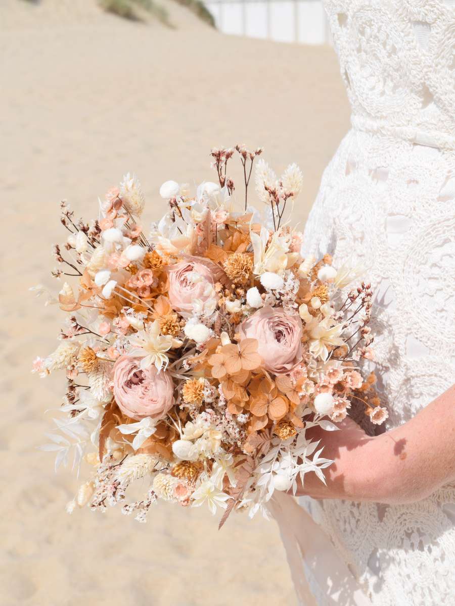 bruid poseert aan het strand en houdt een mokkakleurig bruisboeket vast met een ronde vorm en zachtroze gepreserveerde rozen