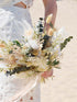 close-up van een modern bruidsboeket van droogbloemen in witte en groene tinten, door een bruid langs haar zijde gehouden