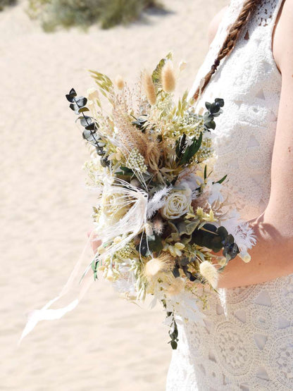 bruid houdt trouwboeket in haar armen vast, gemaakt van droogbloemen in groene, beige en witte tinten