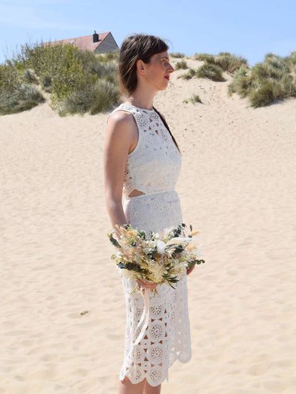 bruid poseert met bruidsjurk en bruidsboeket op het strand en houdt een klein en modern trouwboeket vast met witte en groene droogbloemen zoals eucalyptus, rozen en pampas