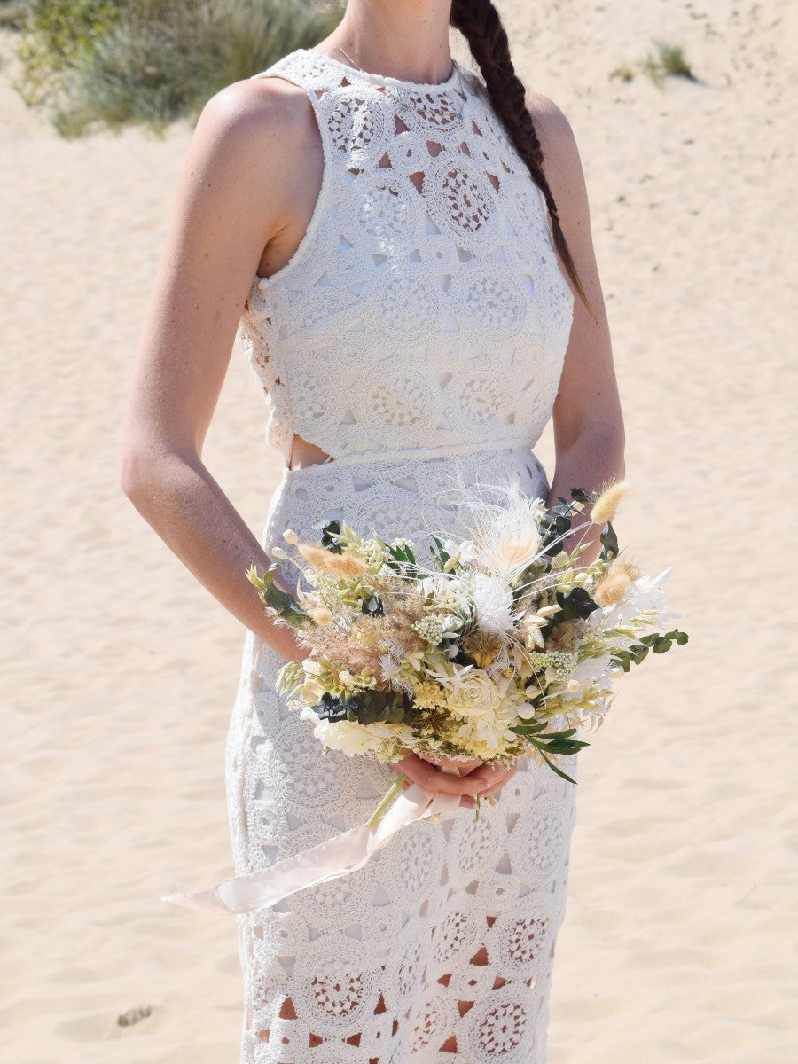 bruid poseert aan de duinen en houdt een modern klein bruidsboeket van droogbloemen vast. Het boeket bevat witte en groene tinten