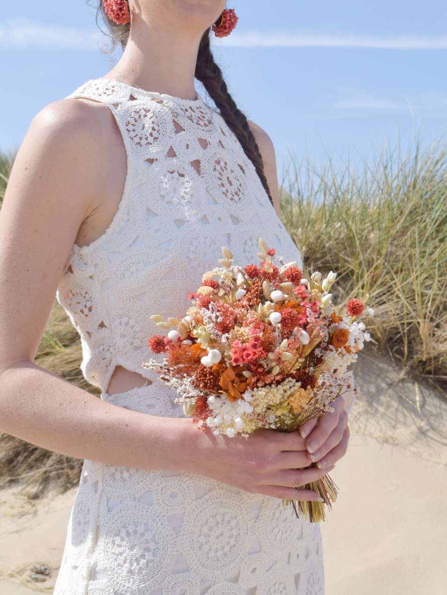 Bruid poseert in de duinen en houdt een klein trouwboeket van droogbloemen in de handen. Het droogboeket is rond, compact en bevat frisse kleuren zoals oranje, terracotta, wit en groen. Ze draagt bijpassende oorbellen van droogbloemen gemaakt