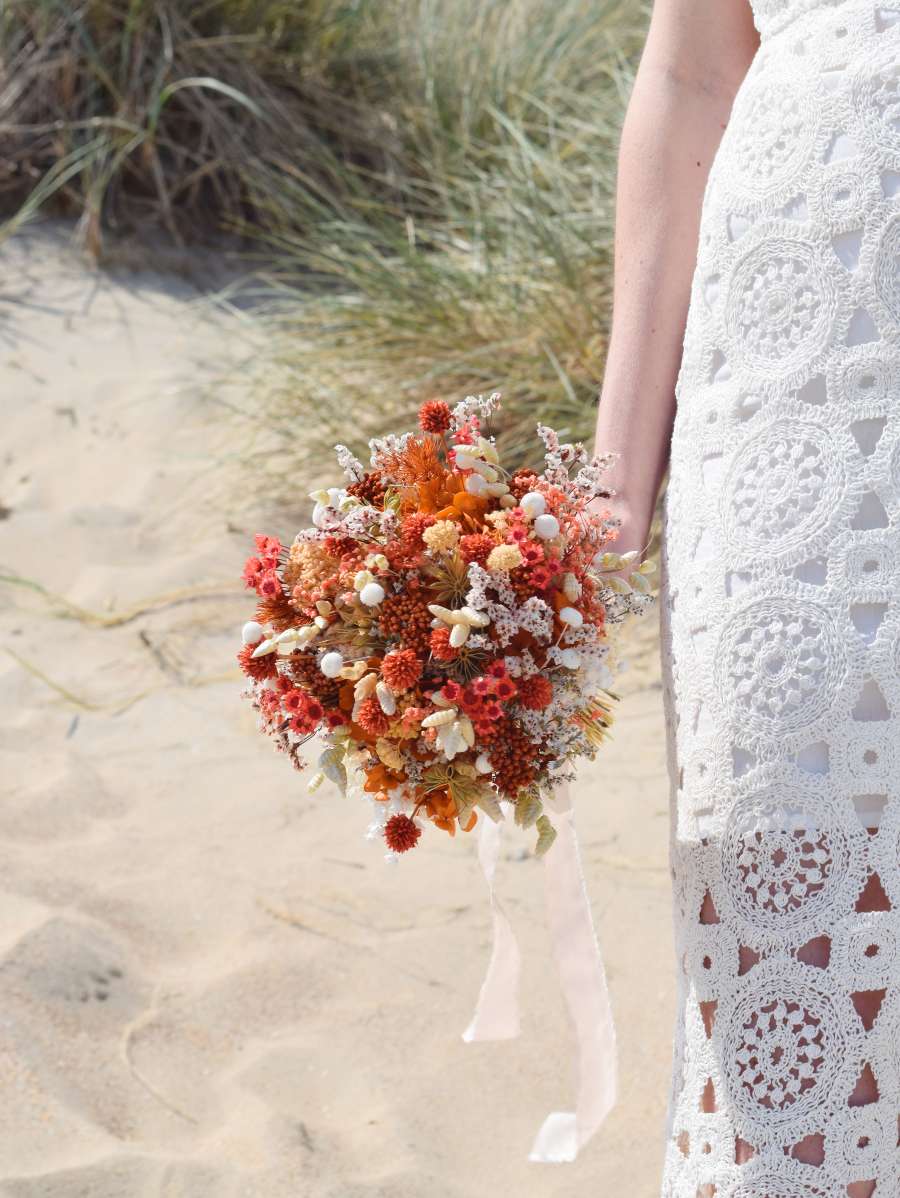 Bruid staat in de duinen en houdt een klein bruidsboeket van droogbloemen langs haar lichaam. Het ronde droogboeket bestaat uit oranje, witte en terracotta tinten en oogt modern