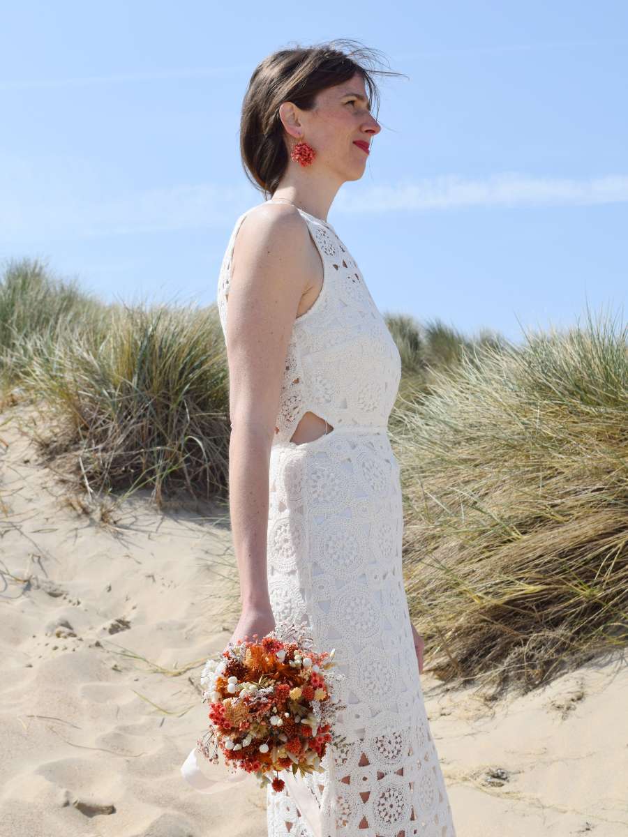 Bruid staat tussen de duinen en laat een klein bruidsboeket van droogbloemen langs haar lichaam hangen. Het droogboeket bestaat uit terracotta, witte en oranje tinten.
