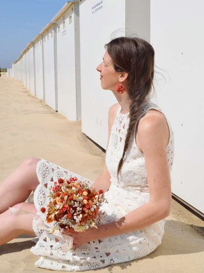 Bruid zit langs strandcabines en houdt een klein bruidsboeket van droogbloemen vast in oranje, witte en terracotta tinten met hortensia, breeza, ruscus en tatarica