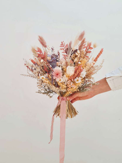 Modern bruidsboeket van droogbloemen wordt in de lucht gehouden en bestaat uit zachtroze, witte, bruine en zachte oranje tinten. Het trouwboeket heeft een lichte X-vorm en bevat ook een witte pauwenveer.