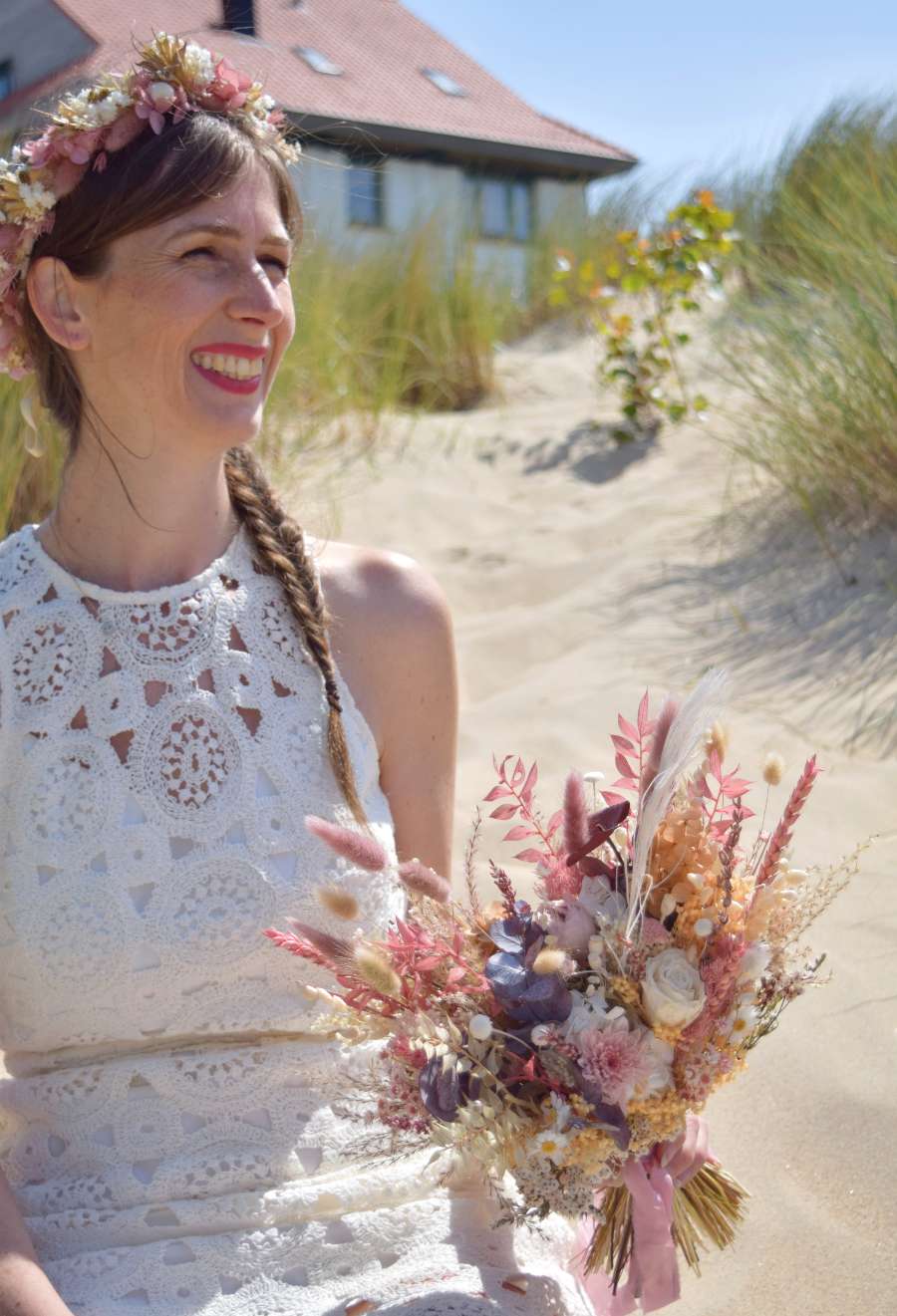 Bruid zit in de duinen en draagt een weelderige roze bloemenkroon met hortensia en heeft in haar handen een modern en X-vormig bruidsboeket van droogbloemen vast in bruine, zachtroze, oranje en witte tinten