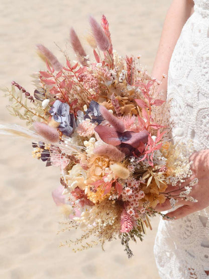 Close-up van bruid die een modern trouwboeket van droogbloemen vasthoudt met bruine eucalyptus, roze bunny tails, witte hortensia en rozen, papavers en oranje broom