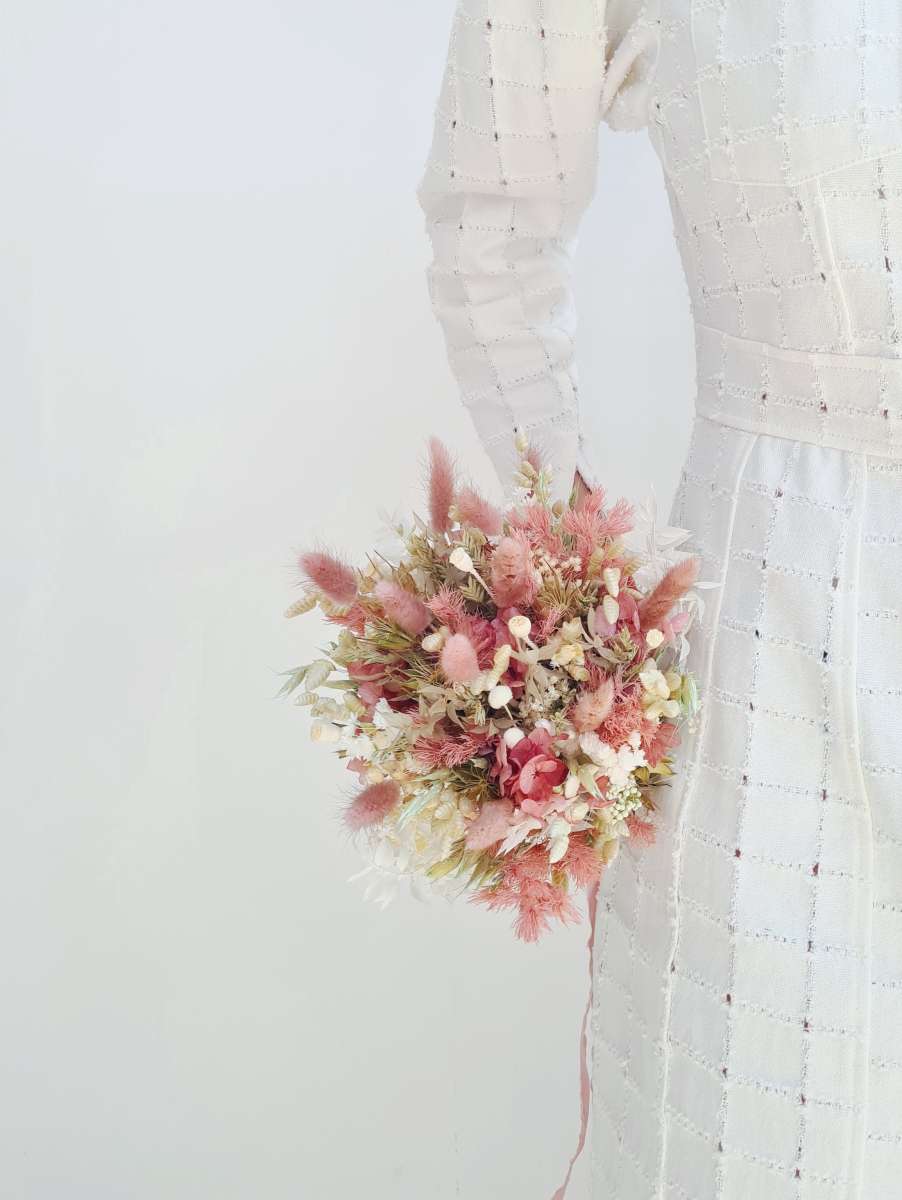 Model houdt klein en rond bruidsboeket van droogbloemen vast in zachtroze en witte tinten.