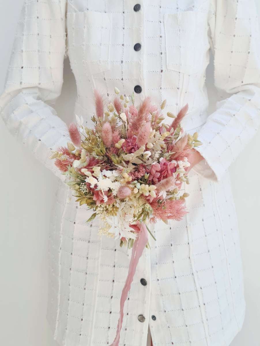 Model houdt een klein bruidboeket van droogbloemen vast met een lang roos lint bevestigd. Het droogboeket bestaat uit zachtroze, witte en groene tinten en oogt romantisch