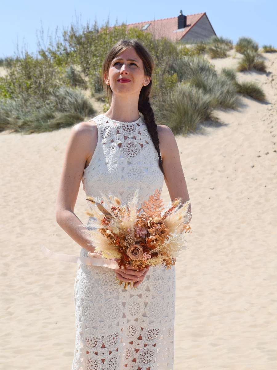 Bruid staat aan de duinen en heeft een modern bruidsboeket gemaakt van droogbloemen vast. Het droogboeket is X-vormig en bestaat uit pampas, mokka gepreserveerde rozen, varens en zoveel meer