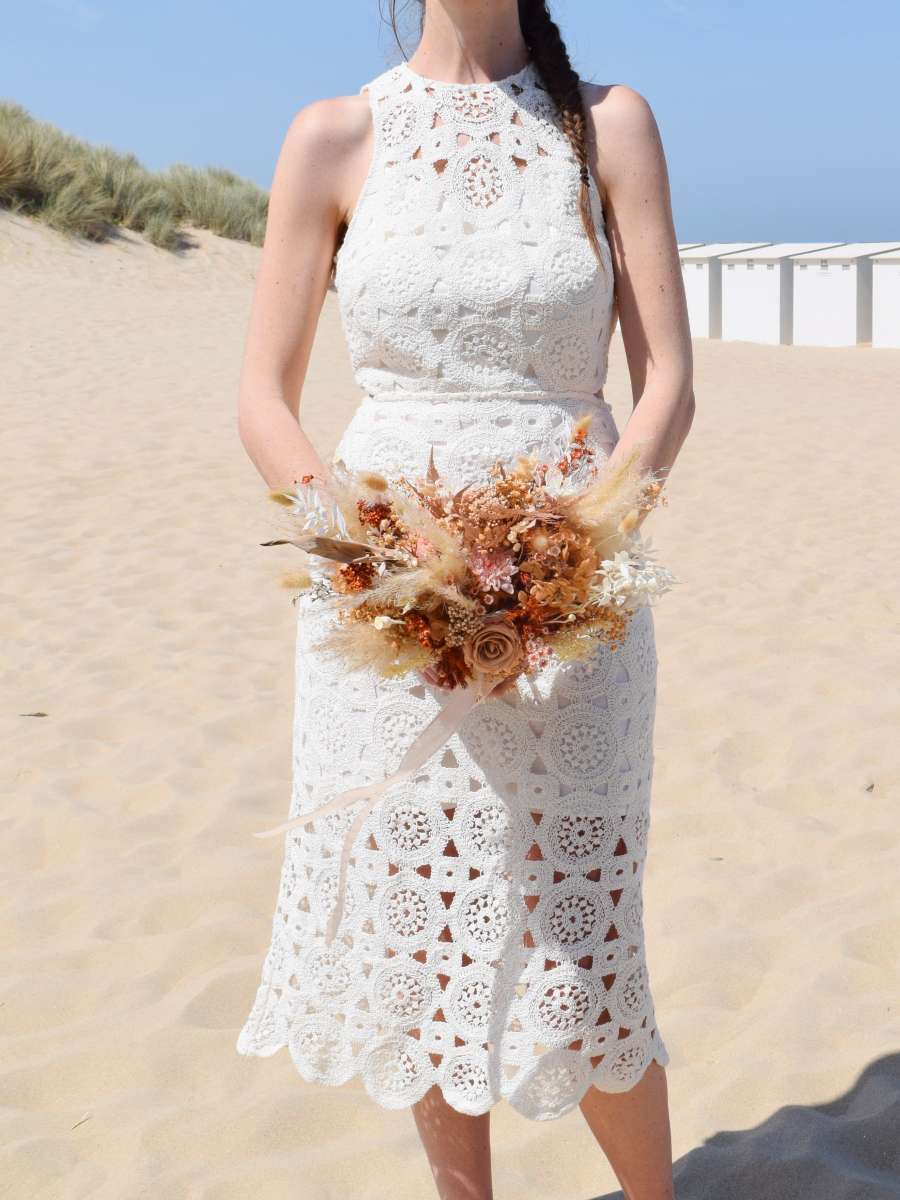 Bruid staat op het strand en draagt een buidsboeket van droogbloemen in natuurlijke tinten met pampas, mokka gepreserveerde rozen, witte ruscus en terracotta rijstbloemen