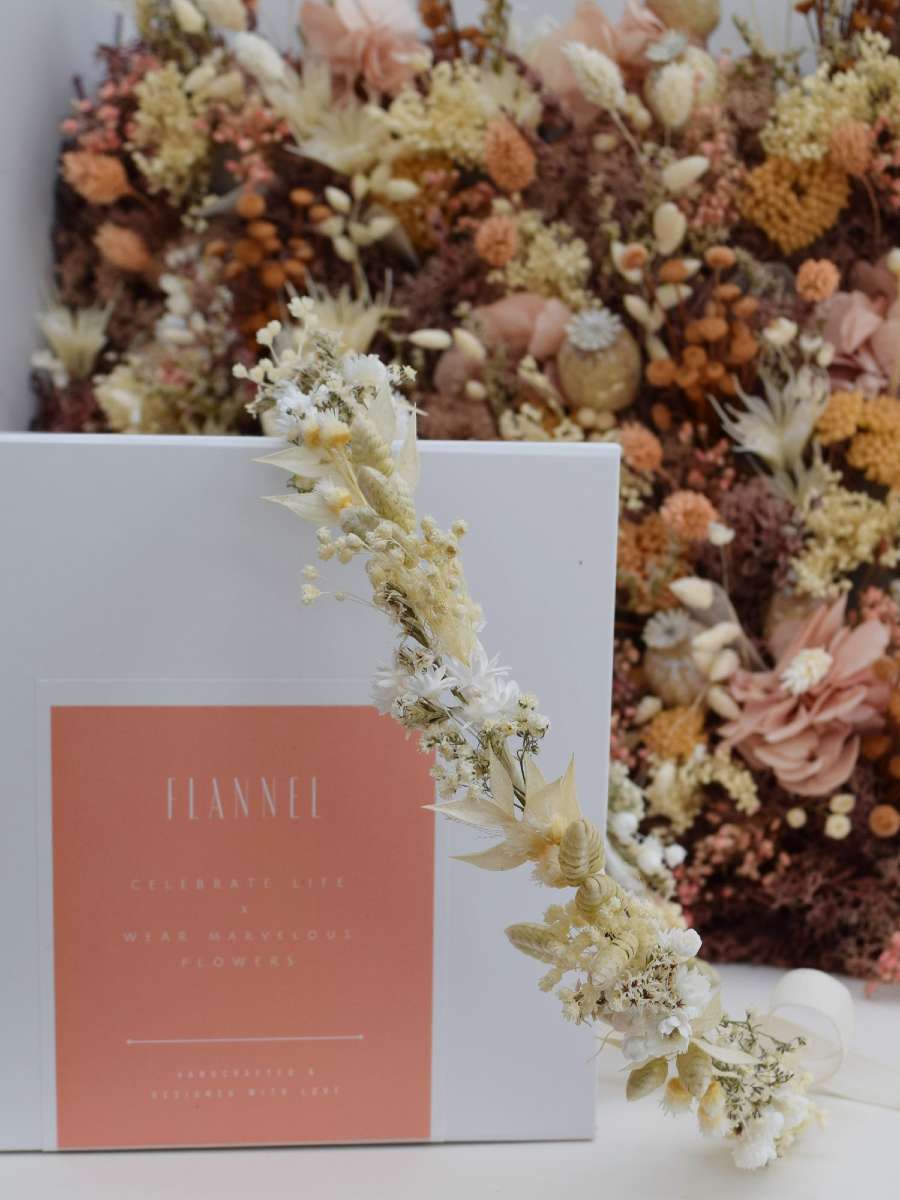 een fijn bloemenkroontje gemaakt van droogbloemen dat over een wit doosje van het merk flannel hangt. Gemaakt van witte, groene en beige droogbloemen
