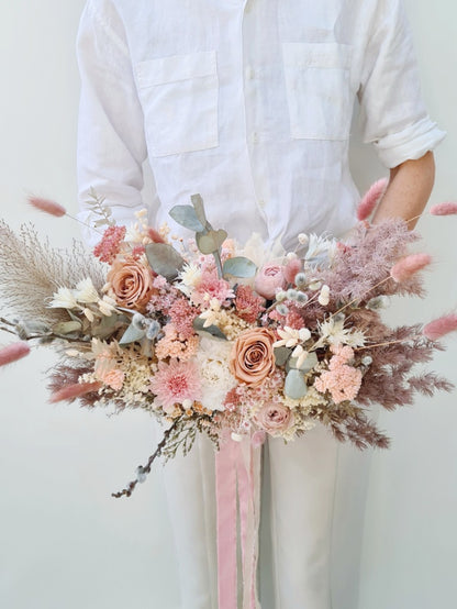 Model houdt groot bruidsboeket van droogbloemen voor zich, opgemaakt met droogbloemen in zachte tinten zoals beige pampas, roze chrysanten, bunny tails en mokka kleurige gepreserveerde rozen